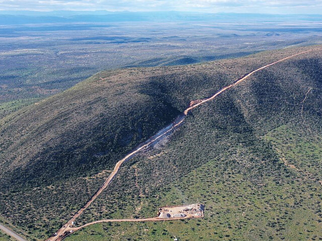 At the bottom of the image, a construction pit is being dug for the foundation of a wind energy plant. In the background, a vast plain stretches out.